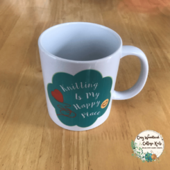 A knitting mug with a turquoise cloud shape with the words "knitting is my happy place" with a ball of yarn in the shape of a heart and a smiley face.